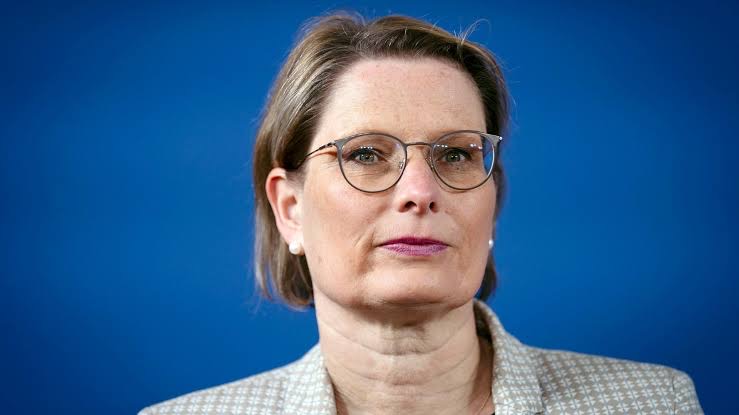 Close portrait of Stefanie Hubig with a serious expression, wearing glasses and a patterned jacket, against a deep blue background.