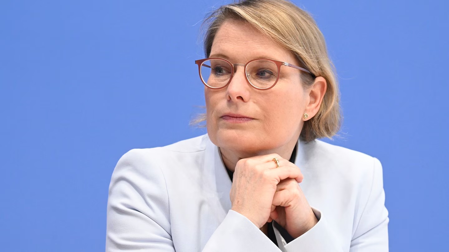 Stefanie Hubig at a press conference in front of a blue backdrop, resting her chin on her folded hands, listening.