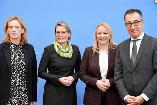 Stefanie Hubig, wearing a green patterned scarf, standing between three colleagues in front of the blue backdrop of the Federal Press Conference in Berlin.