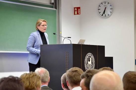 Stefanie Hubig speaking at a lectern in a university lecture hall, with a green chalkboard behind her and an audience in the foreground.