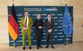 Stefanie Hubig standing between two men in front of a Eurojust backdrop flanked by German and European flags.