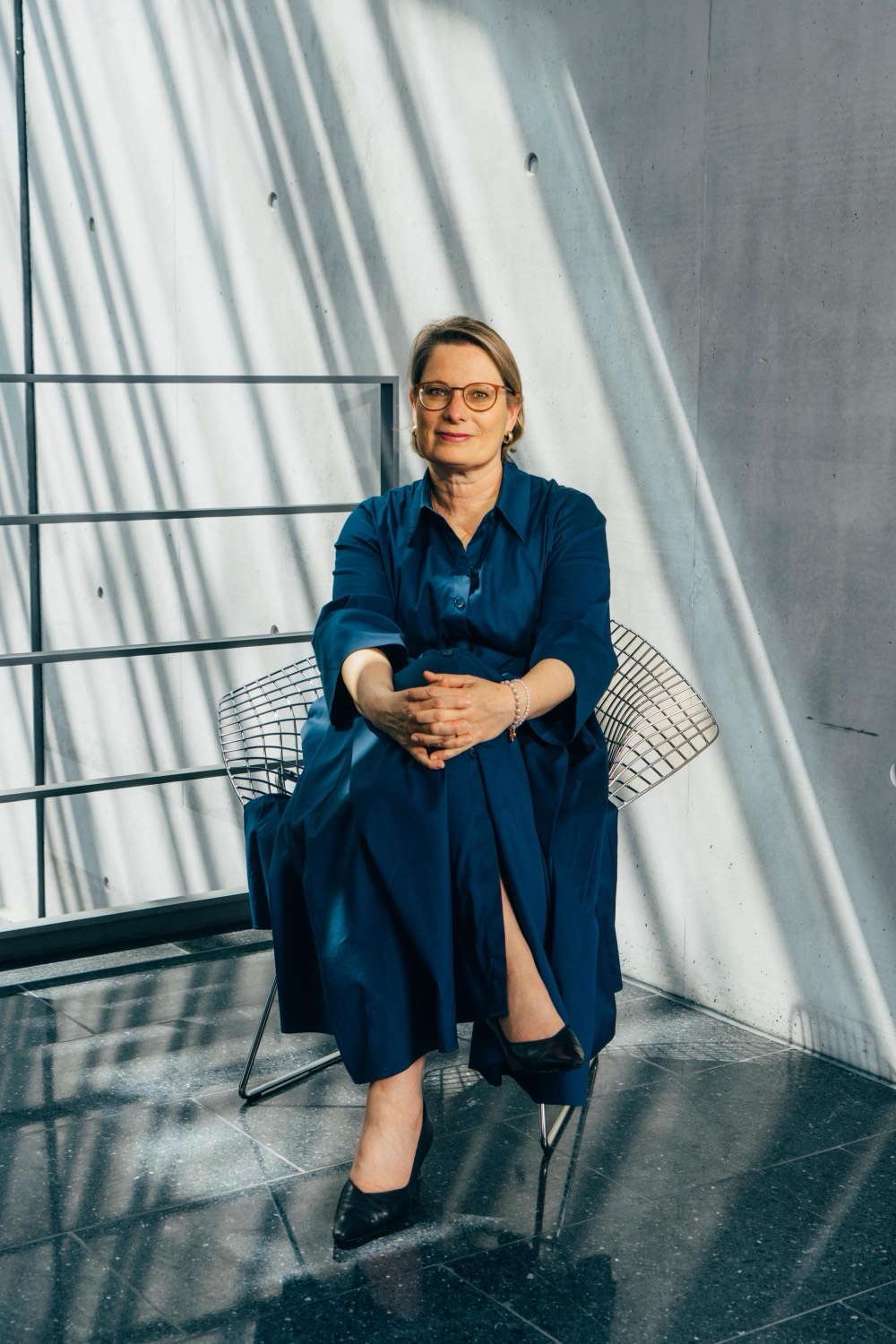 Full-length portrait of Dr. Stefanie Hubig, seated on a wire chair in a sunlit concrete interior, wearing a dark blue dress, hands folded on her knee.