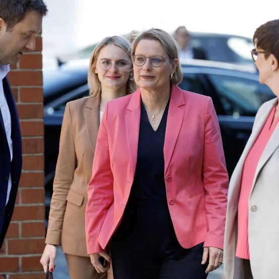 Stefanie Hubig in a coral blazer walking forward among colleagues outside a building.