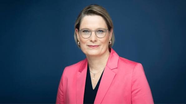 Portrait of Stefanie Hubig smiling, in a rose-pink blazer and glasses, against a dark blue background.