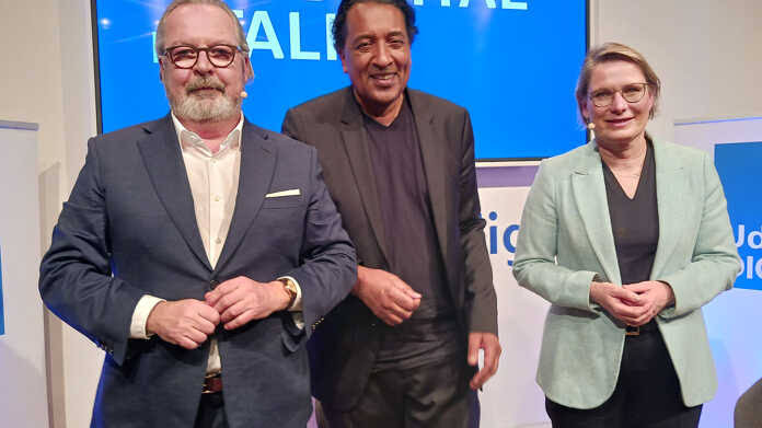Stefanie Hubig in a mint-green blazer standing on a stage with two men after a panel event, in front of a blue screen.