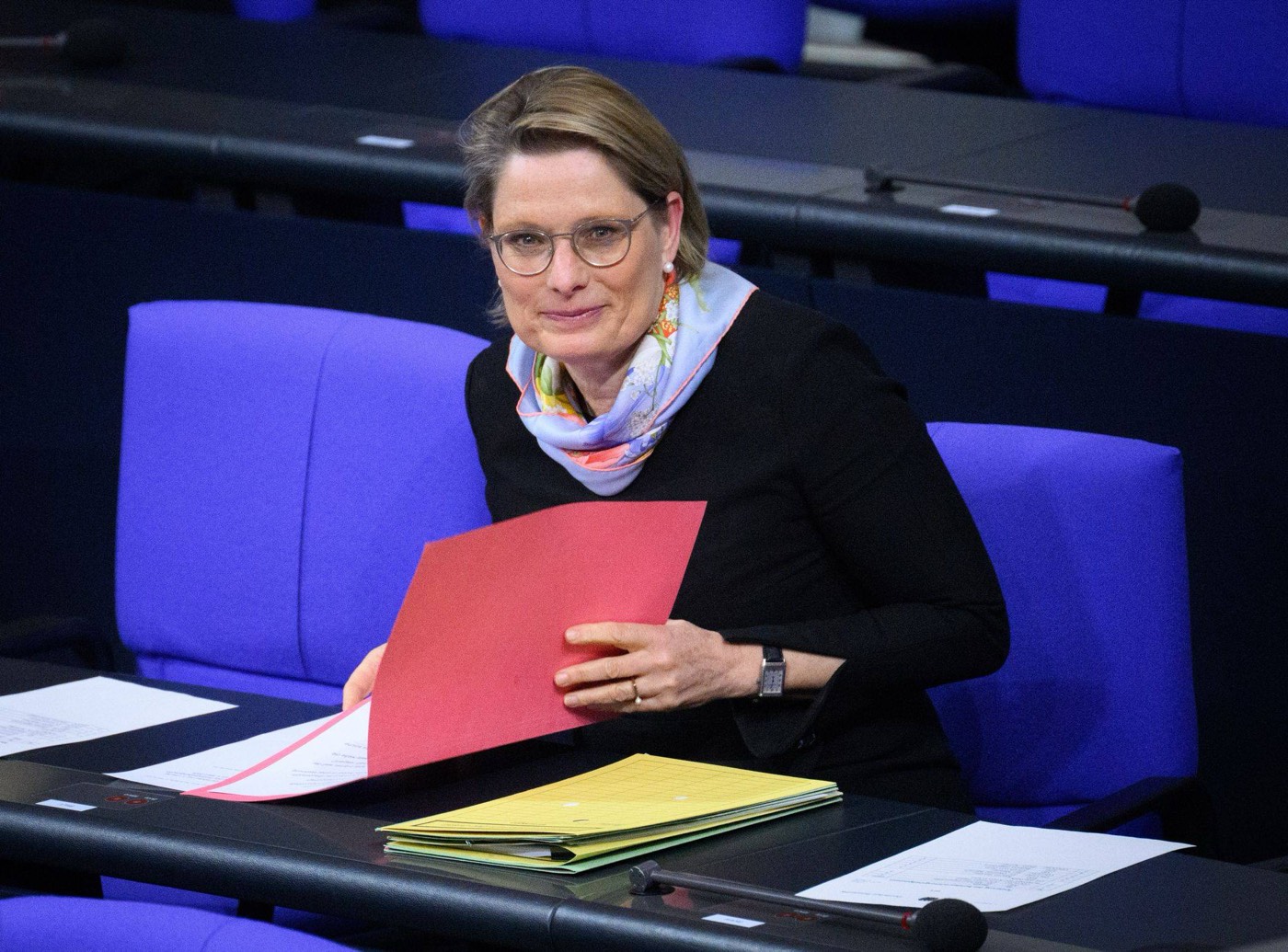 Stefanie Hubig seated on the government bench of the Bundestag, holding a red folder, with yellow folders on the desk in front of her.