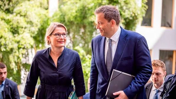 Stefanie Hubig in a dark dress walking and talking with a tall man in a dark suit outdoors in Berlin, with greenery behind them.