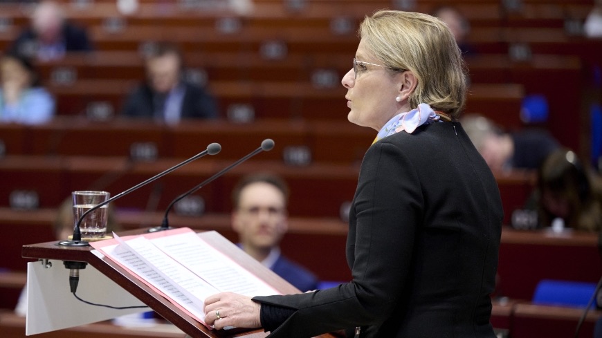 Stefanie Hubig speaking at a lectern with a lavender scarf, in a hemicycle with red seating rows behind her.