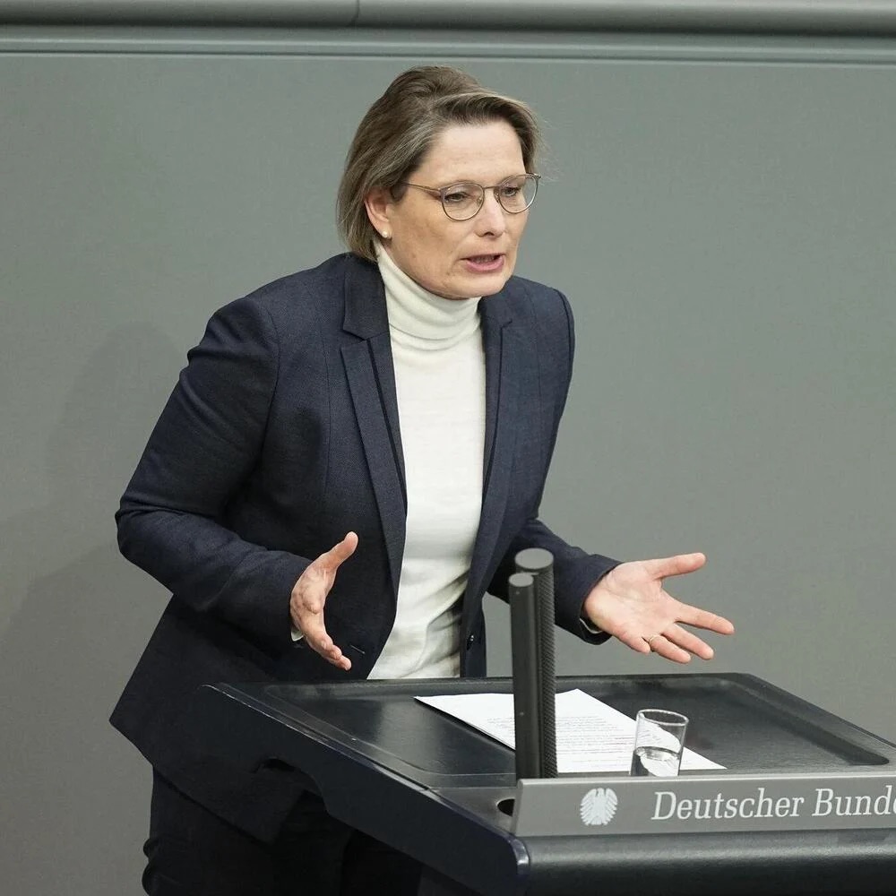 Stefanie Hubig in a navy blazer and white turtleneck speaking at the lectern of the German Bundestag, gesturing with open hands.